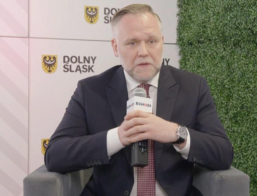 Dawid Jackiewicz przemawiający na wiecu politycznym, z gestykulacją, przy podium z mikrofonem i rozmytym tłumem w tle.