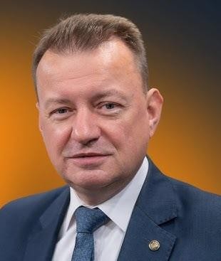 Mariusz Błaszczak, polityk PiS, na tle symboli związanych z bezpieczeństwem i obronnością, reprezentujących jego role jako ministra spraw wewnętrznych i administracji oraz ministra obrony narodowej.