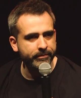 Abelard Giza na scenie z mikrofonem, zamyślony, spoglądający w stronę publiczności, z lekko rozmytym tłem scenicznym.