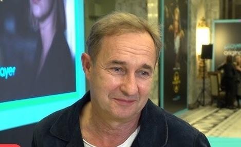 Marek Kalita w roli reżysera, na próbie teatralnej, z aktorami w tle.