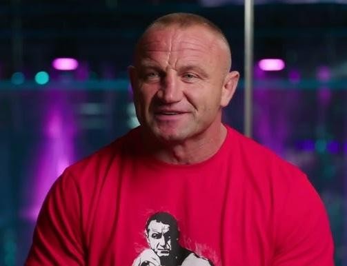 Mariusz Pudzianowski w oktagonie KSW, w dynamicznej pozie bojowej MMA, z widoczną muskulaturą i koncentracją.